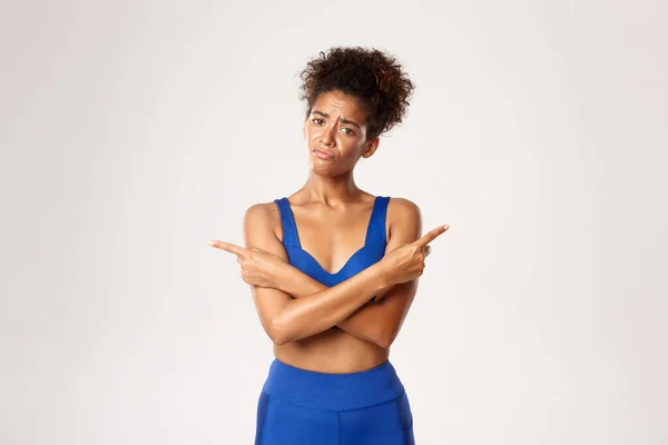 Concept van training en fitness. Triest besluiteloos Afrikaans-Amerikaans meisje in sportkleding, kijken verward, wijzende vingers zijwaarts, witte achtergrond — Stockfoto