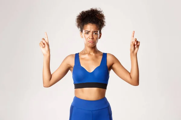 Concept van training en fitness. Aantrekkelijk Afrikaans-Amerikaans sportief meisje in blauwe sportkleding, klagen en kijken verdrietig, wijzende vingers omhoog, witte achtergrond — Stockfoto