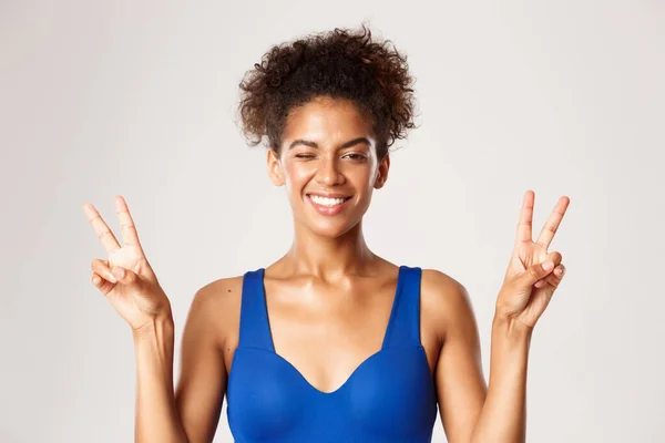 Glad afrikansk-amerikansk fitness flicka i blå träningskläder, visar fred tecken och leende glad, står över vit bakgrund — Stockfoto