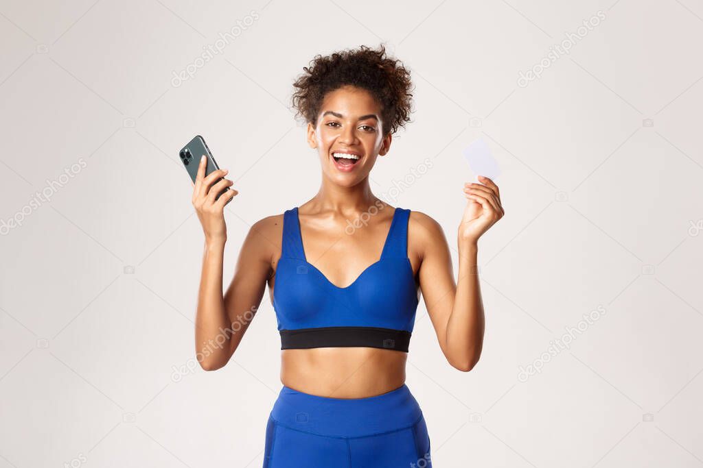 Cheerful african-american female athlete in sport outfit, rejoicing and smiling, holding smartphone and credit card, standing over white background