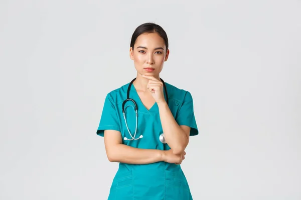 Covid-19, pracownicy służby zdrowia, koncepcja pandemii. Przemyślana lekarka, azjatycka pielęgniarka w fartuchu wyglądająca ciekawie przed kamerą, myśląca, szukająca rozwiązania, stojąca na białym tle rozmyślająca — Zdjęcie stockowe