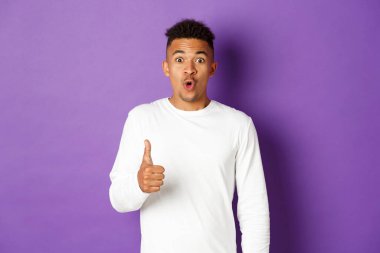 Afro-Amerikan hayranı bir adamın portresi, başparmağını kaldırıyor ve heyecanlı görünüyor, iyi bir ürünü tavsiye ediyor ve övüyor, mor arka planda duruyor.