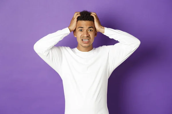 Imagem de homem afro-americano bonito em pânico, de mãos dadas na cabeça e olhando nervoso para a câmera, de pé alarmado sobre fundo roxo — Fotografia de Stock