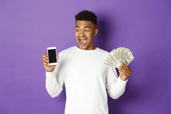 Retrato del hombre afroamericano moderno, mostrando dinero y pantalla del teléfono móvil, sonriendo sorprendido, de pie sobre el fondo púrpura — Foto de Stock