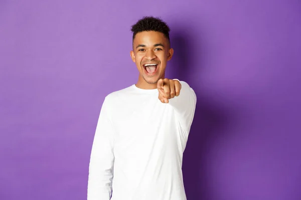 Immagine di un bel giovane afro-americano in felpa bianca, che punta il dito verso la macchina fotografica e sorride, scegliendo te, in piedi su sfondo viola — Foto Stock