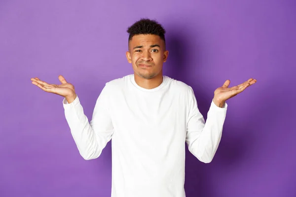 Portrait d'un homme afro-américain indécis en sweat-shirt blanc, levez les mains latéralement et haussant les épaules sans indice, debout confus sur fond violet — Photo