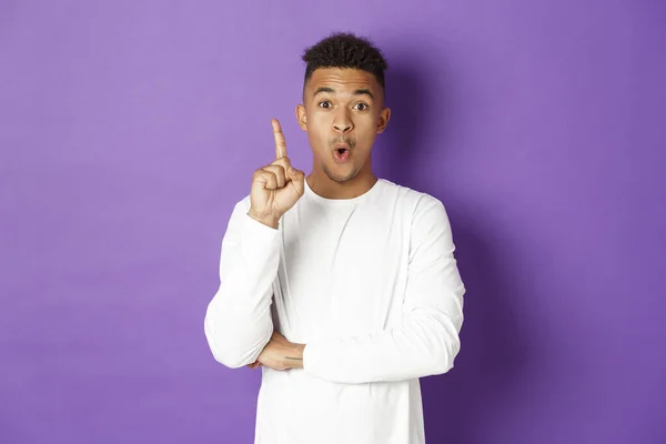 Image of young african-american guy having an idea, raising finger in eureka gesture, suggest plan, think-up solution, standing over purple background — Stock Photo, Image
