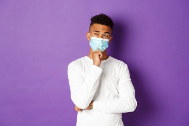 Koronavirüs, karantina ve yaşam tarzı kavramı. Tıbbi maskeli düşünceli bir Afro-Amerikan adamın sol üst köşeye bakıp düşündüğü ya da seçim yaptığı, mor arka planda durduğu görüntüsü.