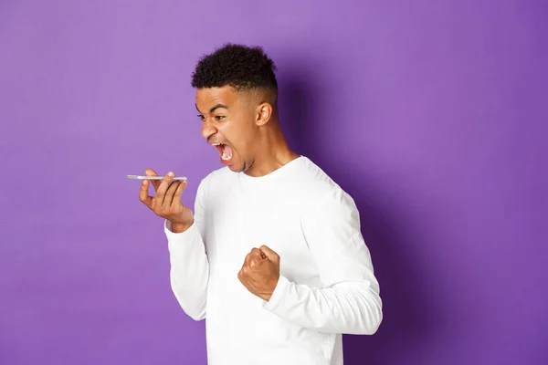 Image of angry african-american man shouting at speakerphone, looking angry and pissed-off, standing with mobile phone over purple background — ストック写真