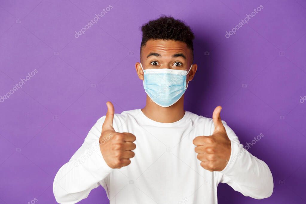 Concept of coronavirus, quarantine and social distancing. Close-up of young african-american guy in medical mask, looking excited and recommending something, showing thumbs-up in approval
