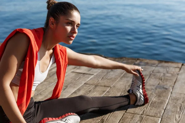 Foto al aire libre del atractivo calentamiento de la mujer fitness antes de correr, sentado en el muelle y estirando las piernas — Foto de Stock