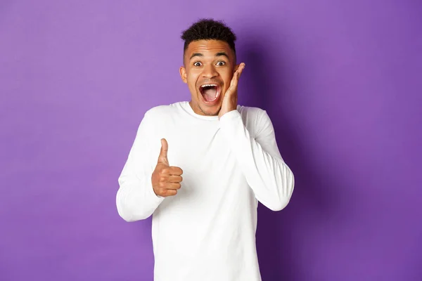 Retrato de homem afro-americano maravilhado, mostrando polegares para cima e olhando animado, recomendar e elogiar bom produto, de pé sobre fundo roxo — Fotografia de Stock