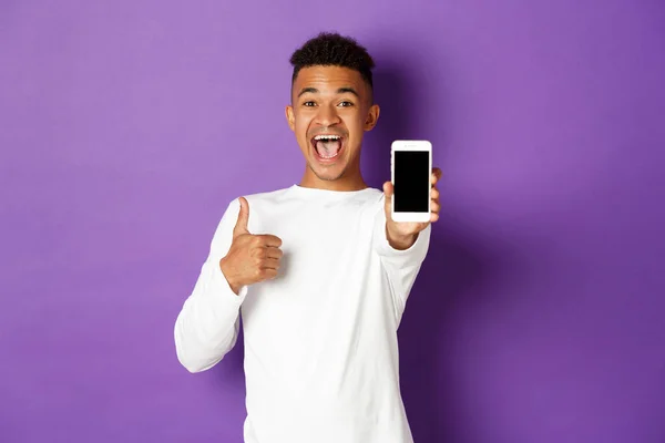 Beyaz eşofmanlı Afro-Amerikan bir adamın resmi, gülümseyen ve onaylanmış başparmaklarını gösteren, cep telefonu ekranı gösteren, mor arka planda duran — Stok fotoğraf
