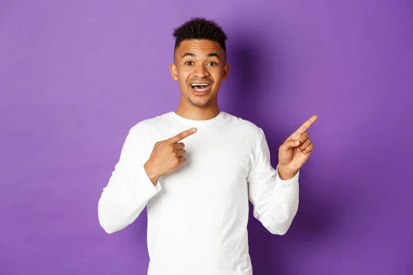 Bild eines glücklichen afrikanisch-amerikanischen männlichen Modells in weißem Sweatshirt, mit den Fingern auf die rechte obere Ecke zeigend und lächelnd, Promo zeigend, auf lila Hintergrund stehend — Stockfoto
