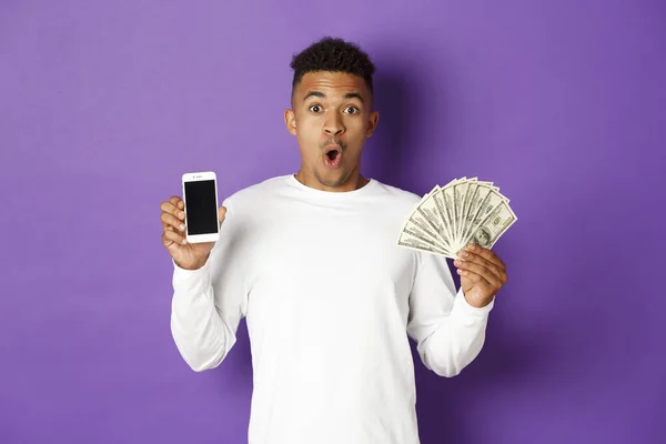 Retrato de hombre afroamericano guapo en sudadera blanca, mostrando el dinero y la pantalla del teléfono móvil, mirando sorprendido, de pie sobre fondo púrpura — Foto de Stock