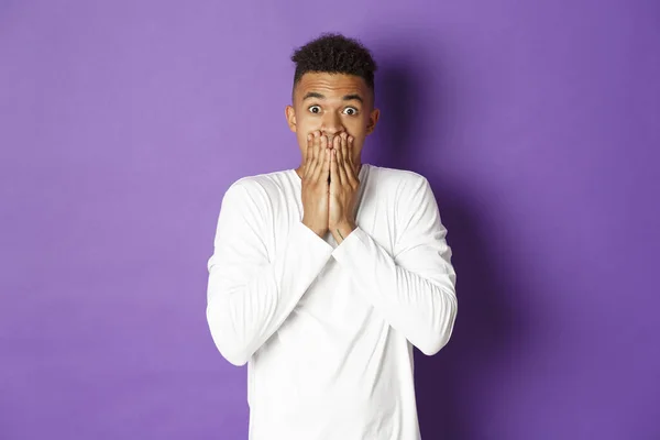 Image of shocked african-american hipster guy in white sweatshirt, gasping and looking startled, cover mouth with hands, standing over purple background — Stock Photo, Image