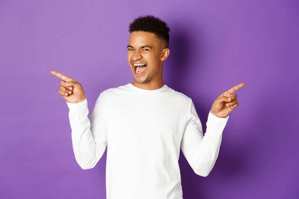 コピースペースで指を横に向け笑顔で製品を証明紫の背景を示す白いスウェットシャツの明るいアフリカ系アメリカ人の男のイメージ — ストック写真