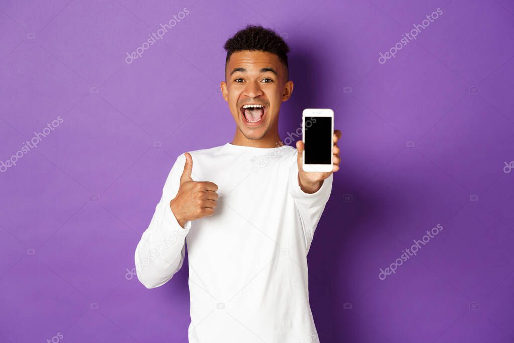 Image of african-american guy in white sweatshirt, smiling and showing thumbs-up in approval, showing mobile phone screen, standing over purple background