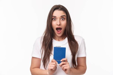 Seyahat, tatil, yaz konsepti. Etkilenmiş, şok olmuş genç bir kadının yakın plan portresi pasaport tutuyor, çenesini indiriyor ve kameraya bakıyor, seyahat etmeye hazır, yurtdışına taşınıyor.