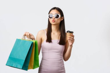 Yaşam tarzı, eğlence ve turizm konsepti. Güneş gözlüklü ve şık Asyalı kadın, kahve içiyor, alışveriş torbaları tutuyor, yaz tatili için yeni kıyafetler alıyor.