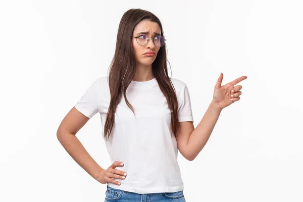 Waist-up portrait of uneasy, sad and gloomy young brunette woman in glasses, looking and pointing finger right with sulking unhappy face, express jealousy or regret, grieving white background — Stock Photo, Image