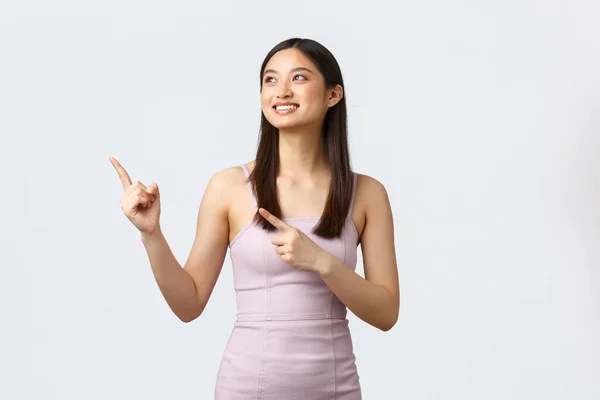 Mujeres de lujo, fiesta y concepto de vacaciones. Feliz mujer asiática sonriente en elegante vestido de noche, estudiante en la noche de graduación mirando esquina superior izquierda, señalando los dedos a la promo tienda, fondo blanco — Foto de Stock