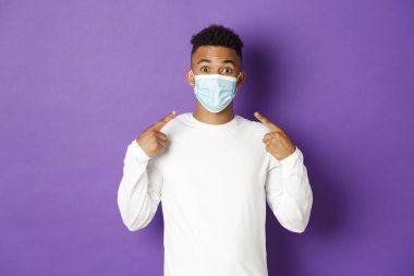 Covid-19 kavramı, salgın hastalık ve sosyalleşme. Afro-Amerikalı bir adamın görüntüsü, Coronavirus sırasında tıbbi maske takmayı öneriyor, mor arka planda duruyor.