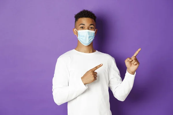 Conceito de coronavírus, quarentena e estilo de vida. Bonito homem afro-americano em máscara médica, apontando os dedos no canto superior direito, mostrando logotipo no espaço de cópia, fundo roxo — Fotografia de Stock