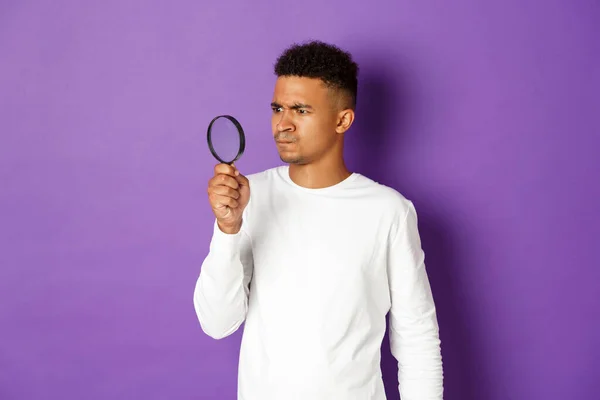 紫色の背景の上に立っている奇妙なものを拡大鏡を通して見て、混乱したアフリカ系アメリカ人の男のイメージ — ストック写真