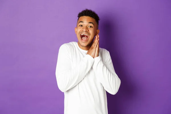 Imagen de chico gay afro-americano emocionado y esperanzado, mirando con anticipación y emoción algo, aplaudiendo manos y sonriendo feliz, de pie sobre el fondo púrpura —  Fotos de Stock