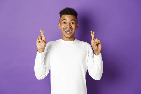 Imagem de jovem afro-americano bonito em camisola branca, olhando esperançoso e sorrindo com os dedos cruzados, fazendo desejo ou esperando por boas notícias, de pé sobre fundo roxo — Fotografia de Stock