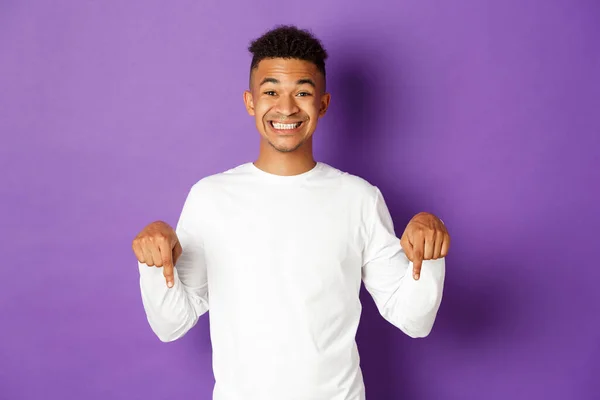 Portrait of african-american happy guy, wearing white sweatshirt, pointing fingers down and smiling cheerful, showing advertisement, standing over purple background — Stock Photo, Image