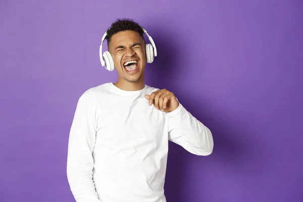 Obrázek bezstarostného afroameričana, jak tancuje, poslouchá hudbu v bezdrátových sluchátkách a zpívá, stojí nad fialovým pozadím — Stock fotografie