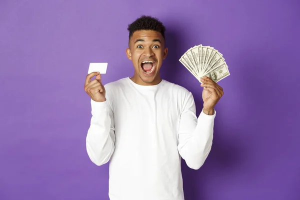 Imagen de chico afroamericano guapo, mirando complacido y sonriente, mostrando tarjeta de crédito y dinero, de pie sobre fondo púrpura — Foto de Stock