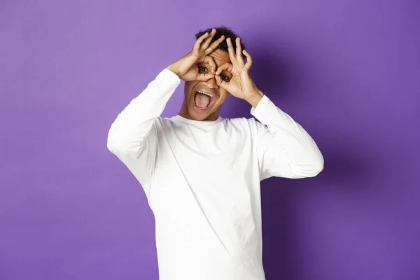Bild von lustigen und glücklichen afrikanisch-amerikanischen Mann in weißem Sweatshirt, Grimassen und Gesichter machen, stehen über lila Hintergrund — Stockfoto