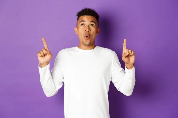 Image of impressed african-american guy in white sweatshirt, pointing and looking up with amazed expression, showing cool advertisement, standing over purple background — Stock Photo, Image