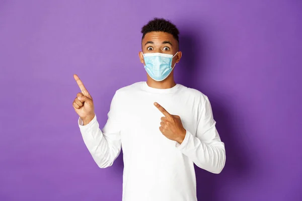 Έννοια του coronavirus, καραντίνα και τρόπος ζωής. Εκπληκτικός αφροαμερικάνος με ιατρική μάσκα που δείχνει διαφήμιση, που δείχνει στην πάνω αριστερή γωνία και χαμογελά, στέκεται πάνω από μωβ φόντο — Φωτογραφία Αρχείου