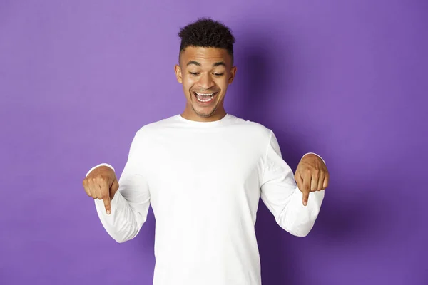 Retrato de animado feliz, homem afro-americano em camisola branca, apontando os dedos para baixo e sorrindo espantado, olhando para a incrível oferta promocional, de pé sobre fundo roxo — Fotografia de Stock