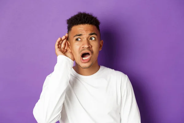 Image de l'homme afro-américain en sweat-shirt blanc écoutant, l'air inquiet, essayant de parler au-dessus de la tête, debout sur fond violet — Photo