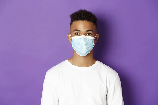 Conceito de coronavírus, quarentena e distanciamento social. Close-up do jovem afro-americano de camisola branca e máscara médica, olhando para a câmera, em pé sobre fundo roxo — Fotografia de Stock