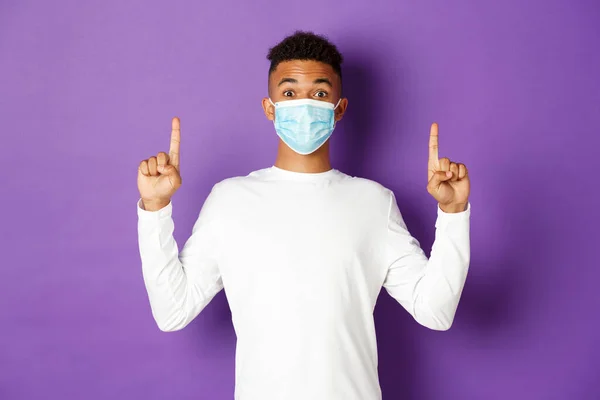 Concept de coronavirus, quarantaine et mode de vie. Homme afro-américain excité en masque médical montrant bannière, pointant les doigts vers le haut à l'espace de copie, debout sur fond violet — Photo