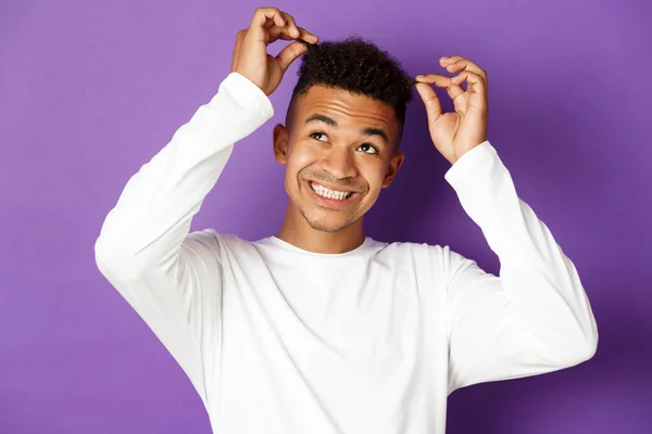 紫色の背景に立って、彼の髪と笑顔に触れて、陽気な若いアフリカ系アメリカ人の男のイメージ — ストック写真