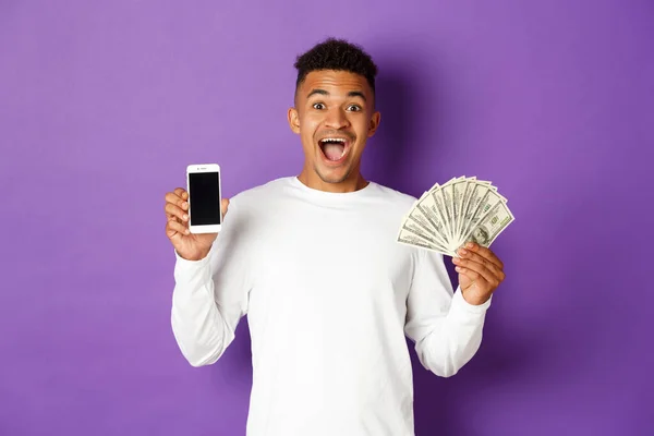 Retrato del hombre afroamericano guapo en sudadera blanca, mostrando el dinero y la pantalla del teléfono inteligente, mirando sorprendido, de pie sobre el fondo púrpura — Foto de Stock