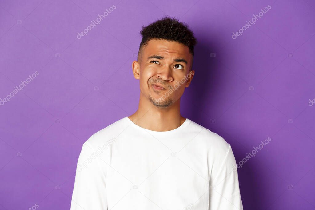 Close-up of indecisive african-american man making choice, looking perplexed at upper left corner, thinking about something complicated, standing over purple background