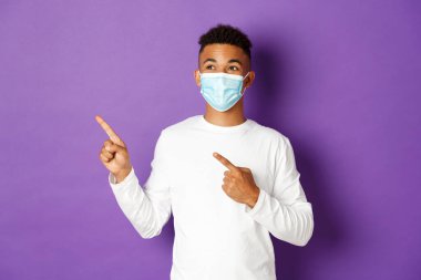 Koronavirüs, karantina ve yaşam tarzı kavramı. Tıbbi maskeli yakışıklı Afro-Amerikan bir adam, sol üst köşeye bakıyor, reklamını yapıyor, mor arka planda poz veriyor.