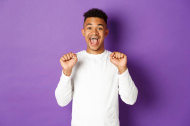 Mutlu ve neşeli Afro-Amerikan beyaz kazak giyen, sevinçten zıplayan, başarıyı ya da başarıyı kutlayan, mor arka planda duran