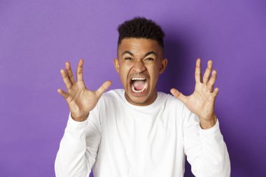 Image of pissed-off african american guy yelling at someone, raising hands and looking with hatred and anger, standing over purple background