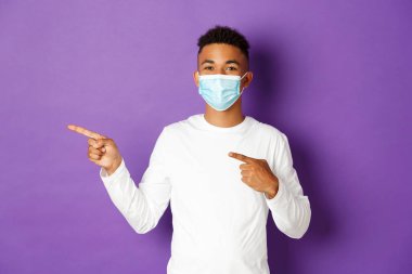 Koronavirüs, karantina ve yaşam tarzı kavramı. Tıbbi maskeli neşeli Afro-Amerikalı adam reklamını yapıyor, solu işaret ediyor ve gülümsüyor, mor arka planda duruyor.
