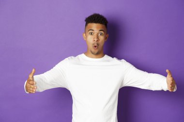 Şaşırmış genç Afro-Amerikalı adam büyük bir şey gösteriyor, geniş bir nesneyi şekillendirmek için ellerini yana açıyor, mor arka planda duruyor.