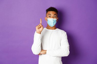 Koronavirüs, karantina ve yaşam tarzı kavramı. Tıbbi maske takan, parmağını kaldıran ve planı öneren Afro-Amerikan genç bir adamın resmi, bir fikri var, mor arka planda duruyor.
